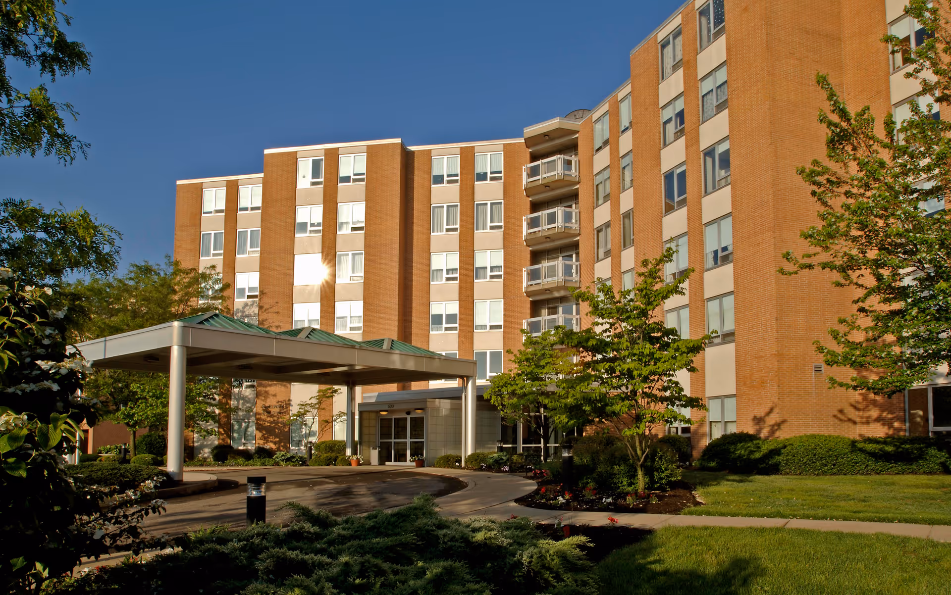 Exterior view of a multi-story brick building with numerous windows, a covered entrance with a green roof, surrounded by green trees and landscaping under a clear blue sky.