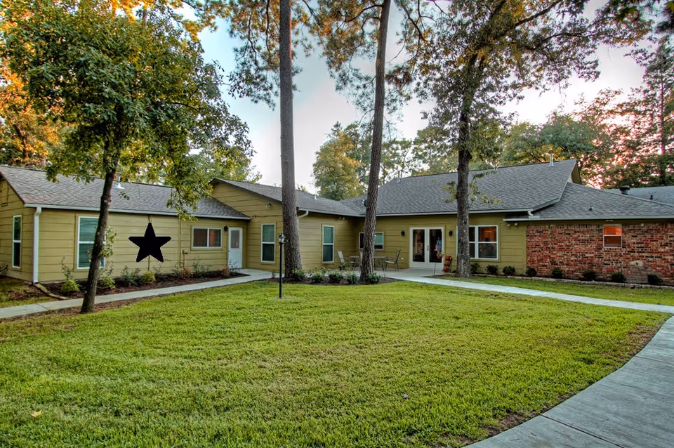 Exterior view of a single-story senior living facility building named The Pines at Oakhurst, featuring a well-maintained green lawn, several tall trees, and a paved walkway leading to the entrance. The building has yellow siding with a section of red brick and multiple windows.