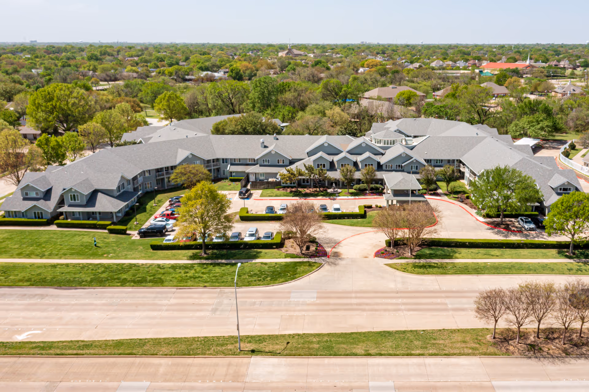 Aerial view of Solstice Senior Living at Plano, showing a large, U-shaped building surrounded by green lawns, trees, and parking areas. The building has a gray roof and multiple windows, with a driveway and entrance area visible at the front.