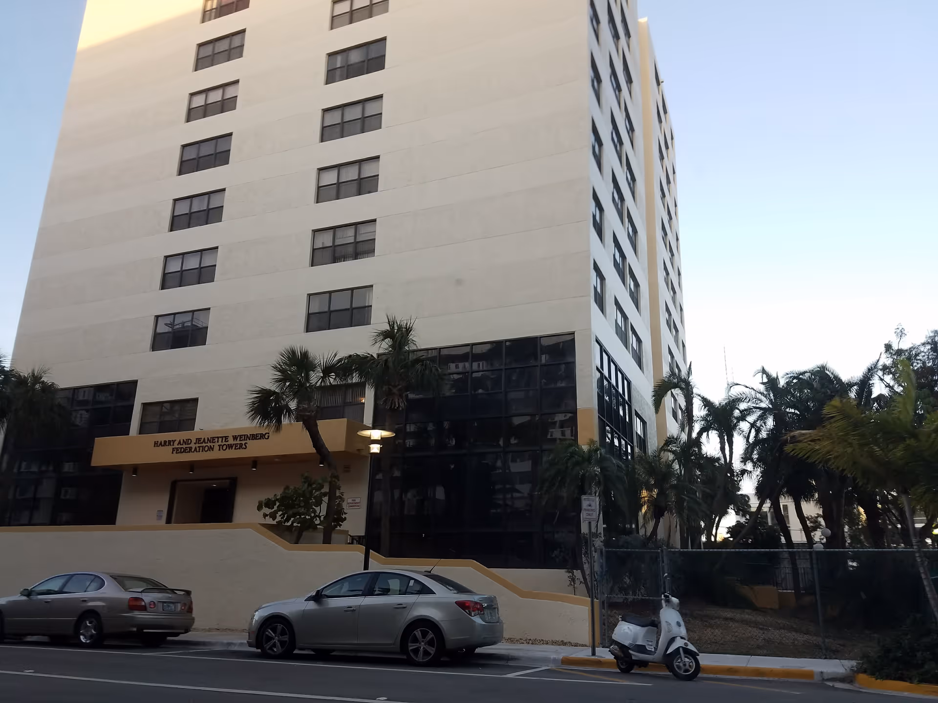 Tall white senior living tower with entrance sign and parked cars outside.