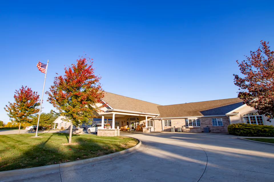 Exterior view of a single-story senior living facility building with a covered entrance, surrounded by green grass and trees with autumn-colored leaves. An American flag is flying on a flagpole near the driveway under a clear blue sky.