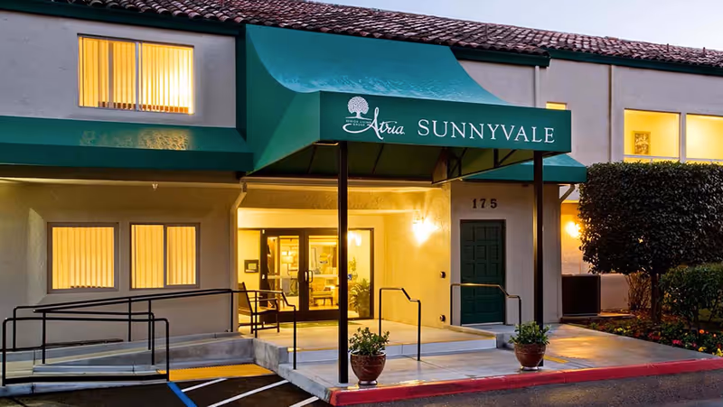 Entrance of Atria Sunnyvale with a green awning, lit windows, double glass doors and an accessibility ramp.