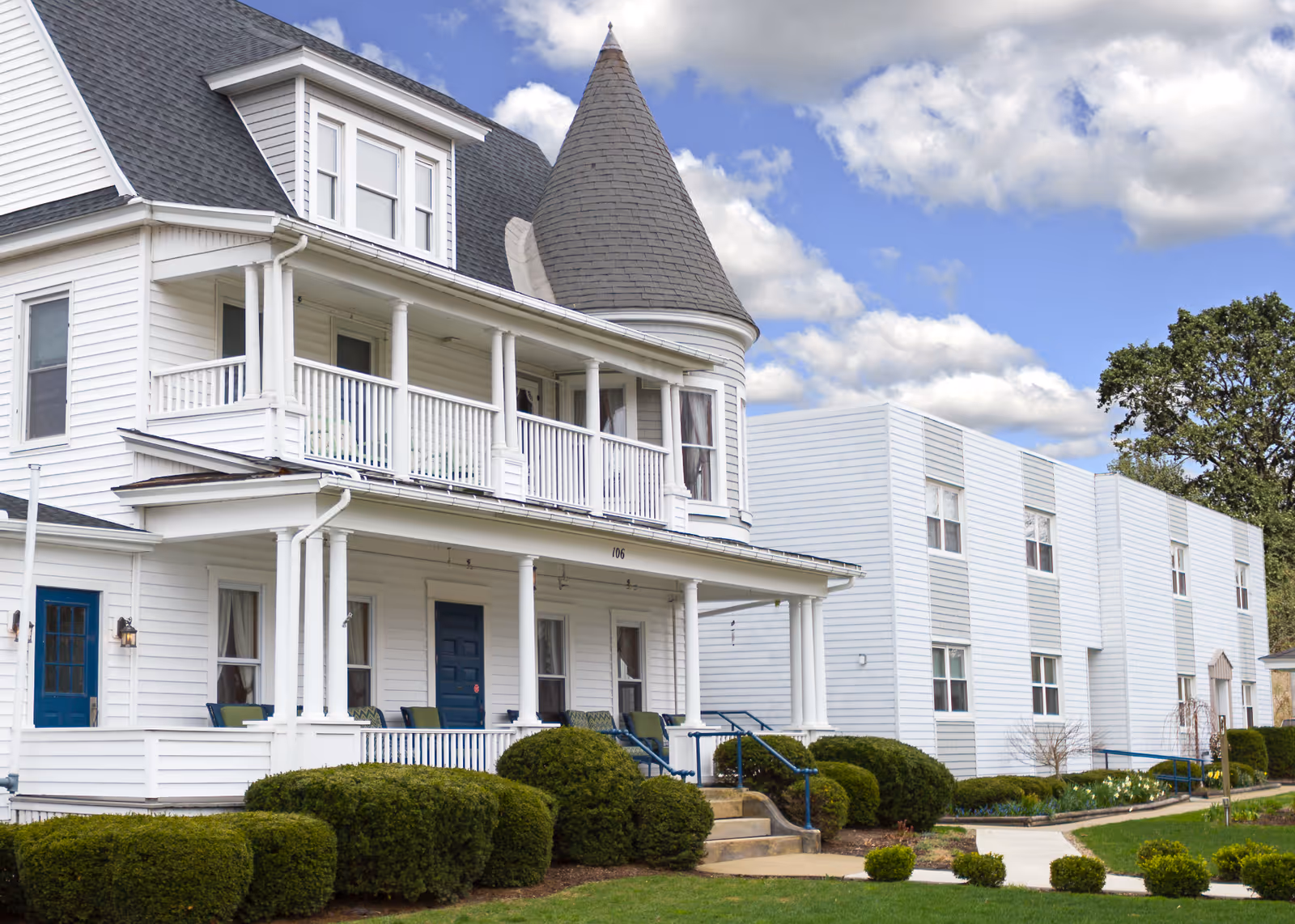 White multi-story senior living residence with porch, turret, and manicured hedges.