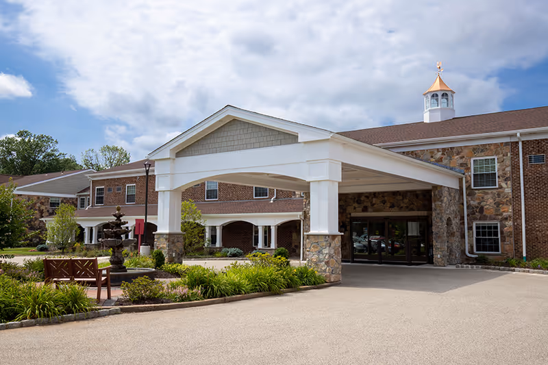 Covered porte-cochere entrance of a brick-and-stone senior living building with a fountain, benches, and landscaped driveway.