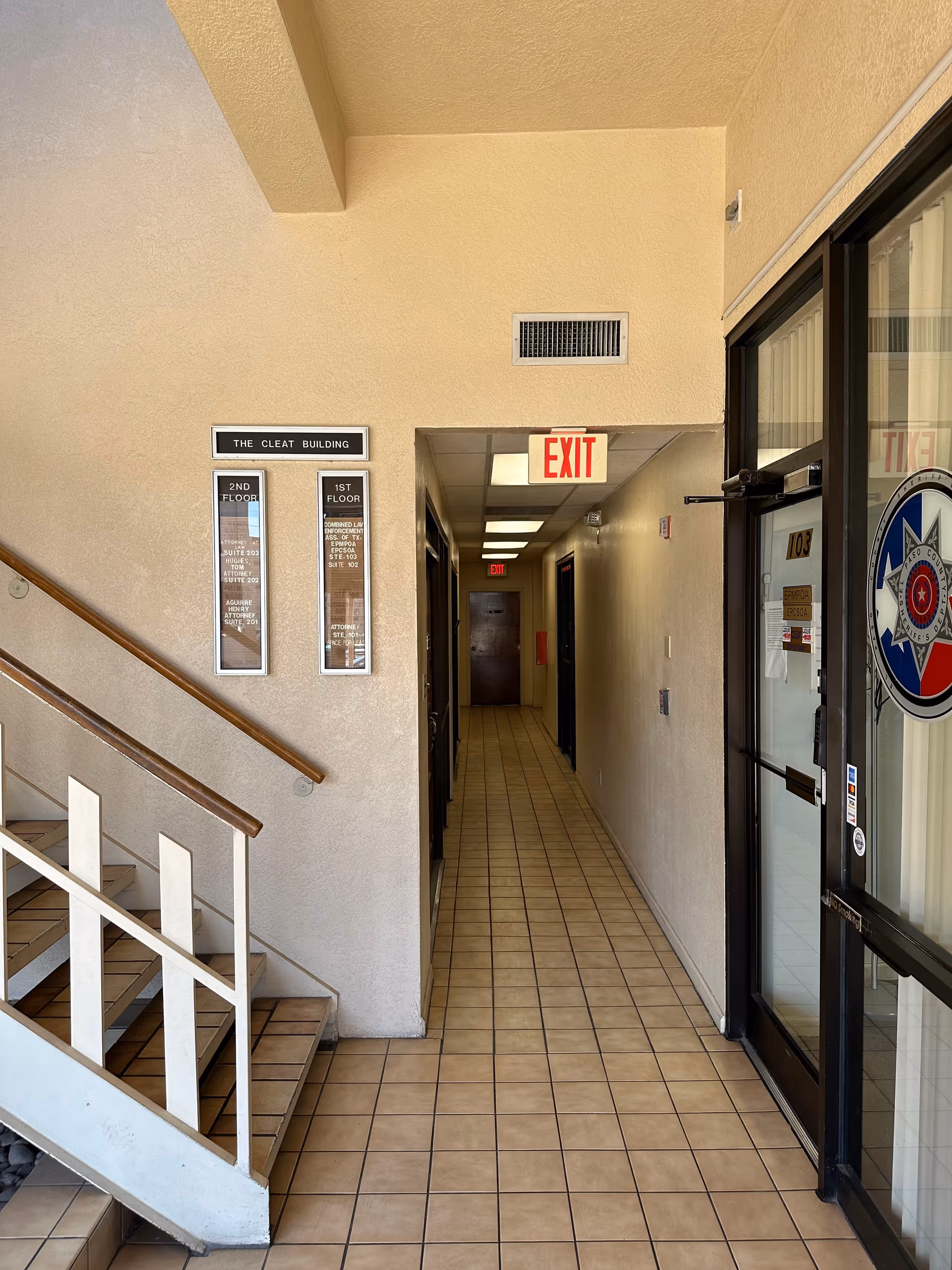 Beige interior hallway with stairs on left, exit sign, and office doors along tiled corridor.