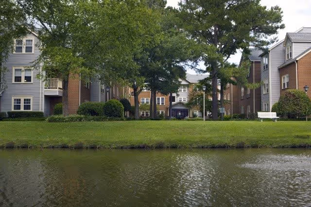 View of a senior living facility named First Colonial Inn showing multiple connected buildings with brick and siding exteriors, surrounded by trees and bushes, with a pond in the foreground and a white bench near the water.