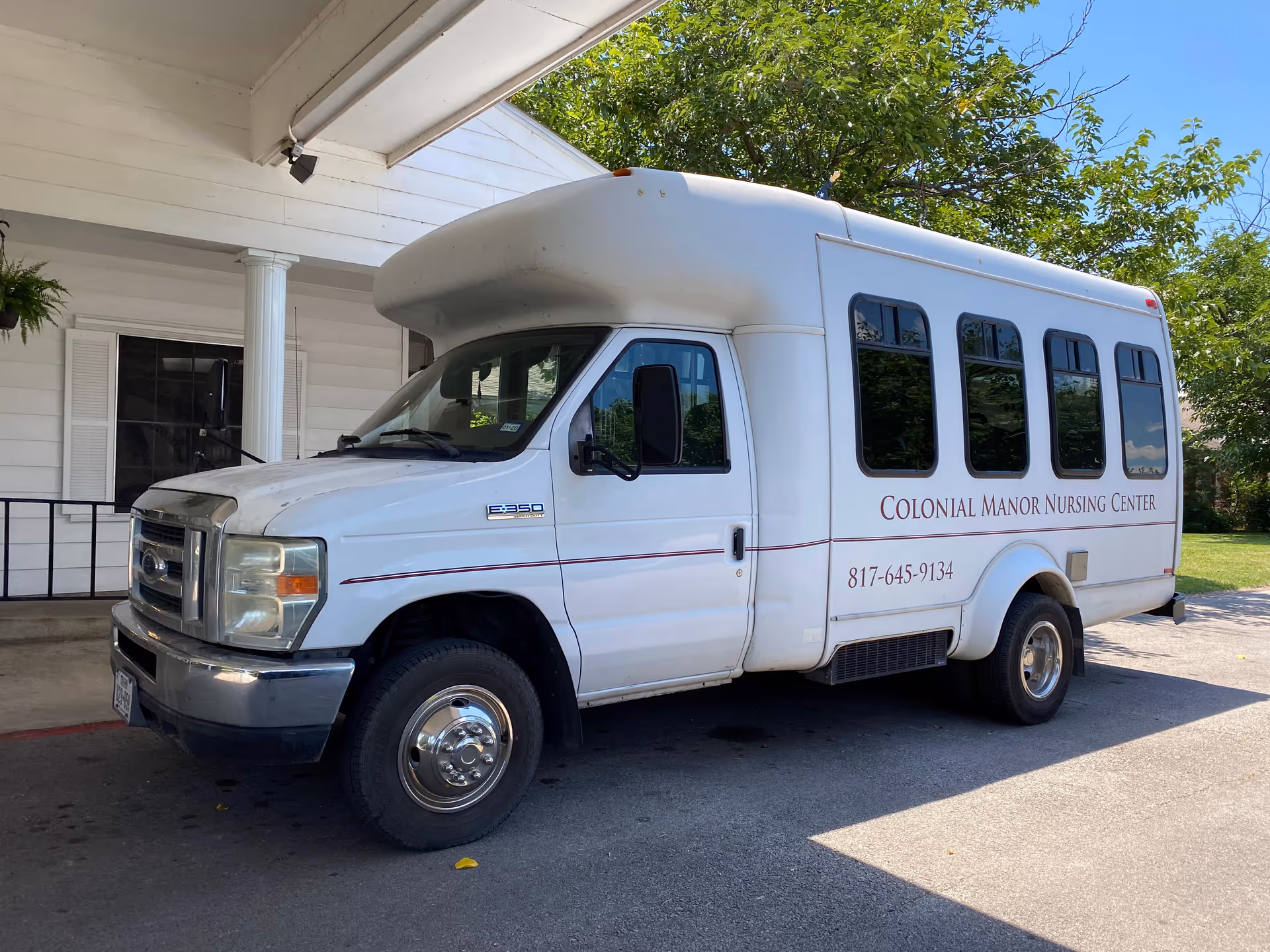 White shuttle van parked under a covered entrance with 'Colonial Manor Nursing Center' and a phone number on its side.