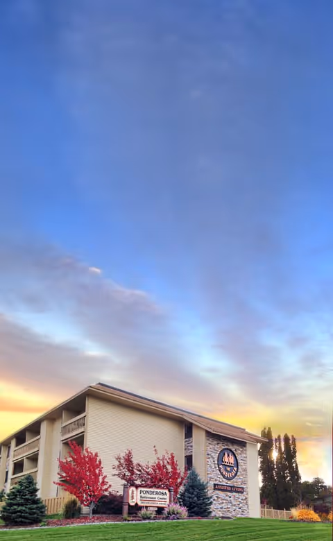 Exterior front of the Ponderosa assisted living building with landscaped lawn, trees, and a large sign beneath a colorful sky.