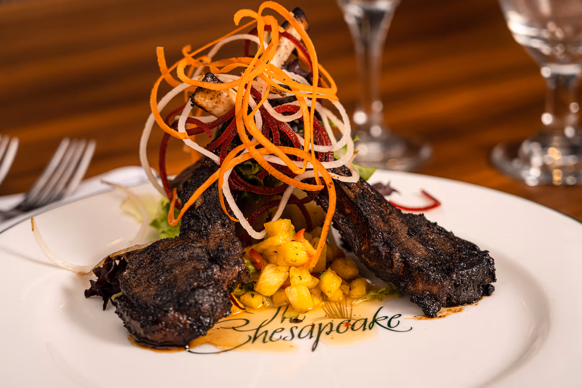 A gourmet dish featuring two grilled lamb chops arranged on a white plate with the word 'Chesapeake' partially visible. The lamb chops are garnished with a colorful spiral of thinly sliced vegetables and served with a small portion of diced yellow fruit or vegetable. The background shows a wooden table with blurred glassware and cutlery.