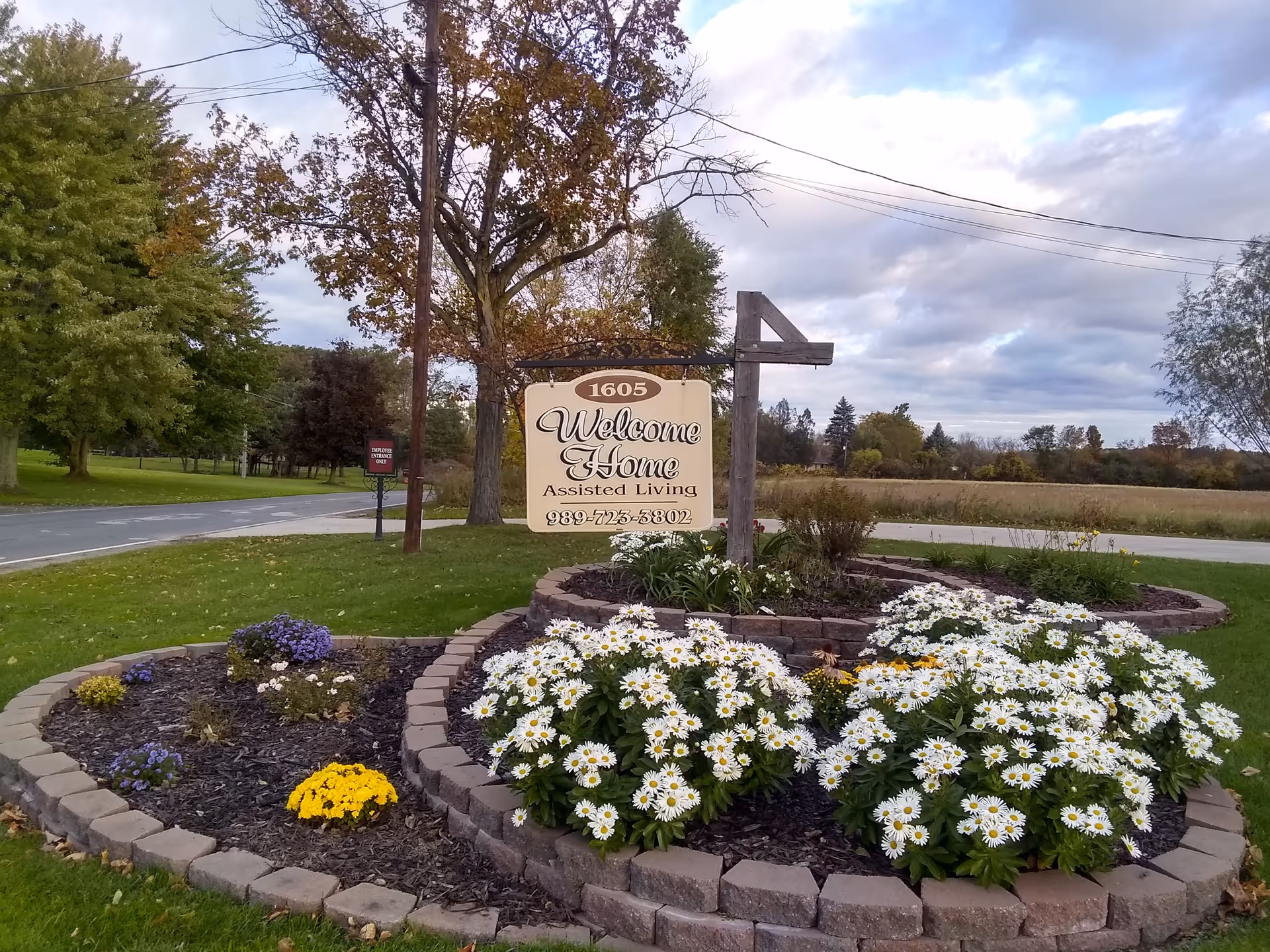 Exterior view of a garden bed with daisies and a Welcome Home Assisted Living sign.
