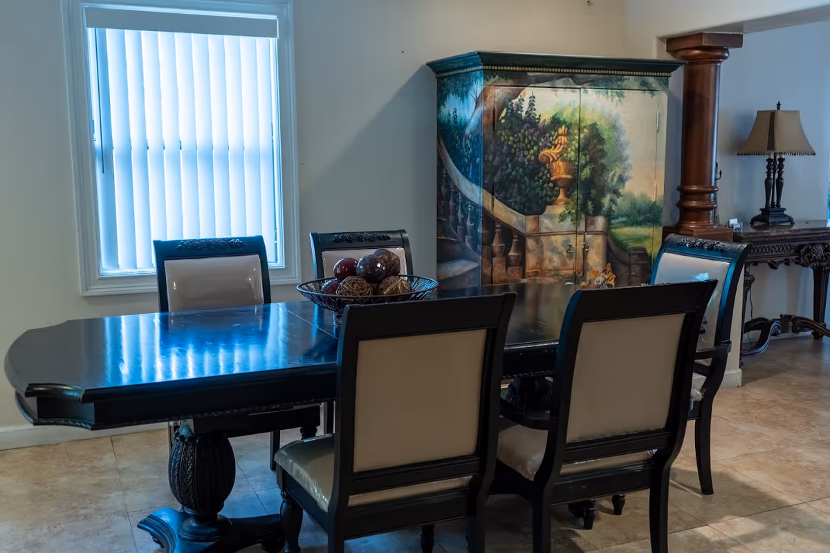 A dining room with a dark wooden table surrounded by six chairs with light-colored upholstery. On the table is a decorative bowl filled with ornamental balls. Behind the table is a painted cabinet featuring a scenic mural of a garden staircase and urn. To the right, there is a wooden column and a side table with a lamp. A window with vertical blinds is on the left wall.