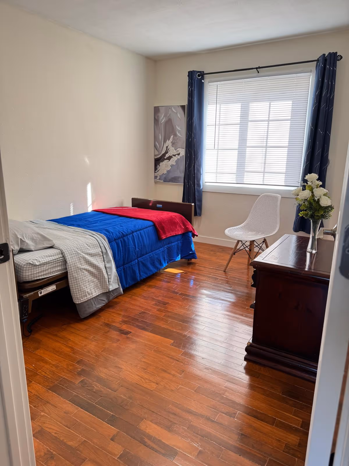 A small bedroom with a bed, blue bedding, window, white chair, and dresser.