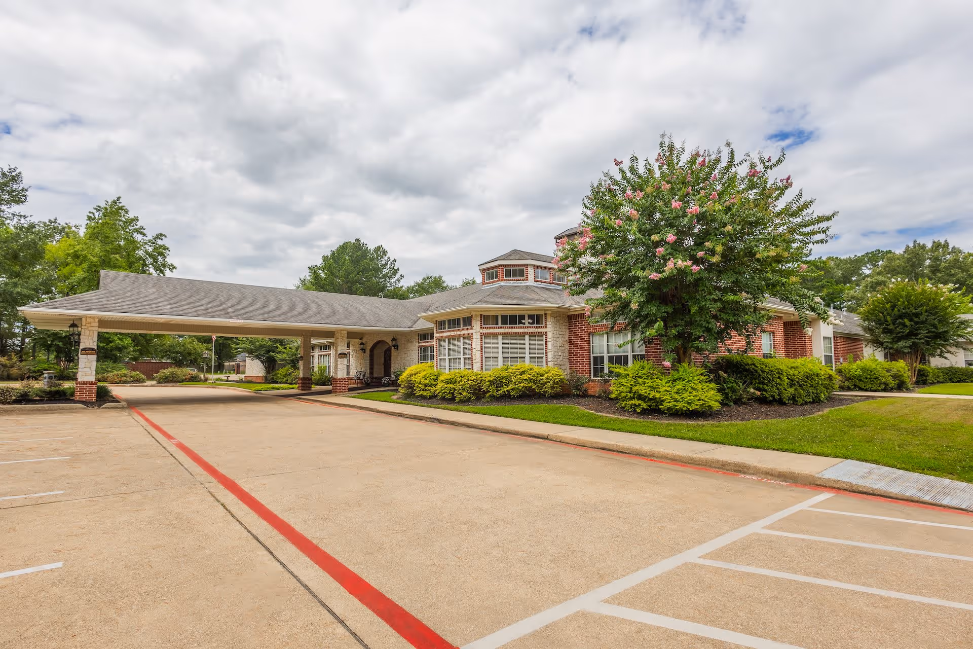 Exterior view of Arabella of Kilgore Senior Living facility showing a covered entrance with brick and stone facade, surrounded by green shrubs and trees under a partly cloudy sky.