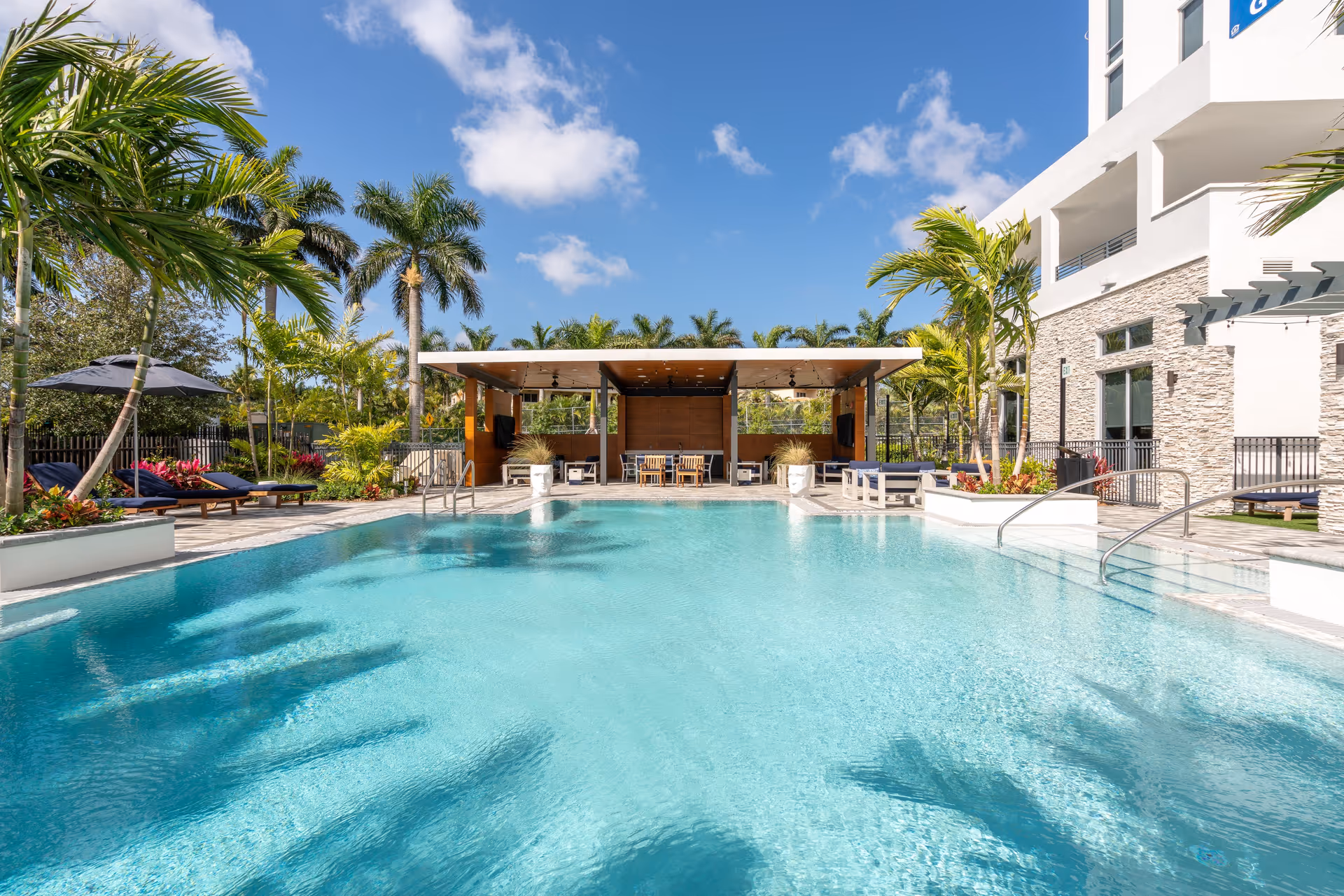 Outdoor swimming pool with lounge chairs, palm trees and a covered cabana beside a modern building under a blue sky.