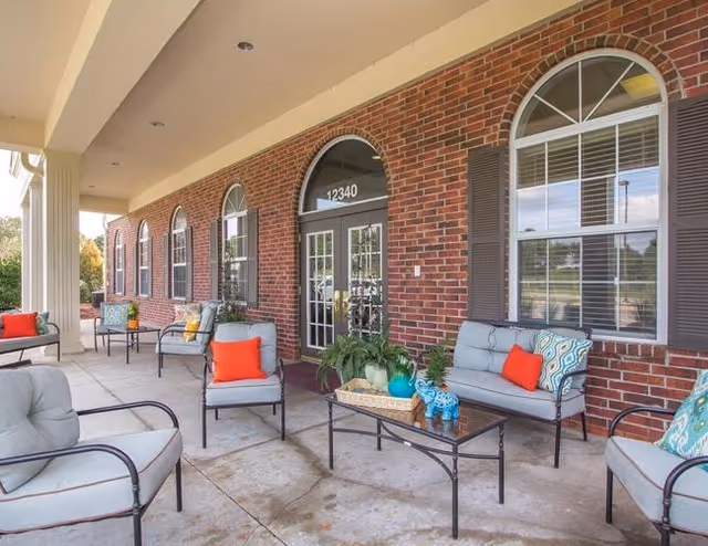 Covered front porch of a brick memory care building with arched windows and outdoor seating and coffee tables.