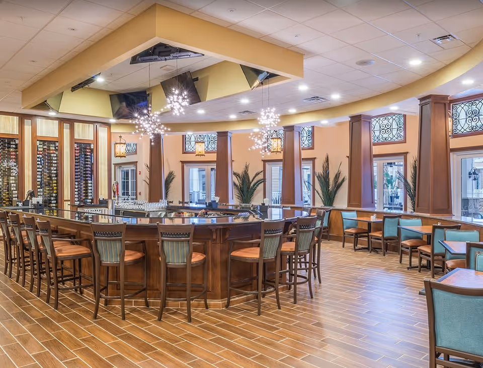 Spacious dining area with a central wooden bar surrounded by stools, tables with teal chairs, decorative hanging lights, and wine racks.