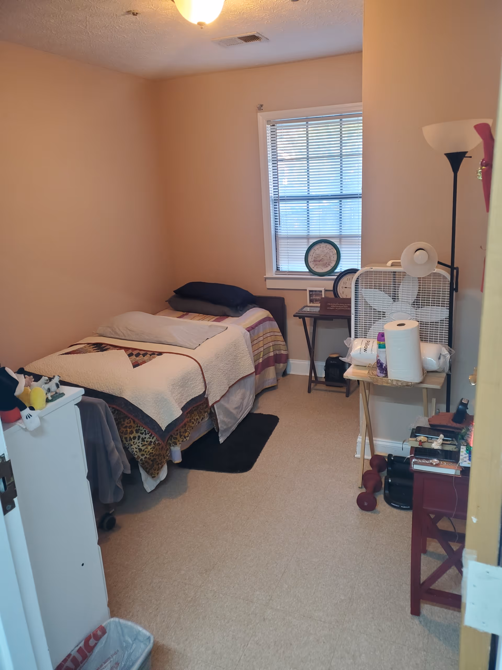 Small bedroom in a senior living facility with bed, fan, and window.