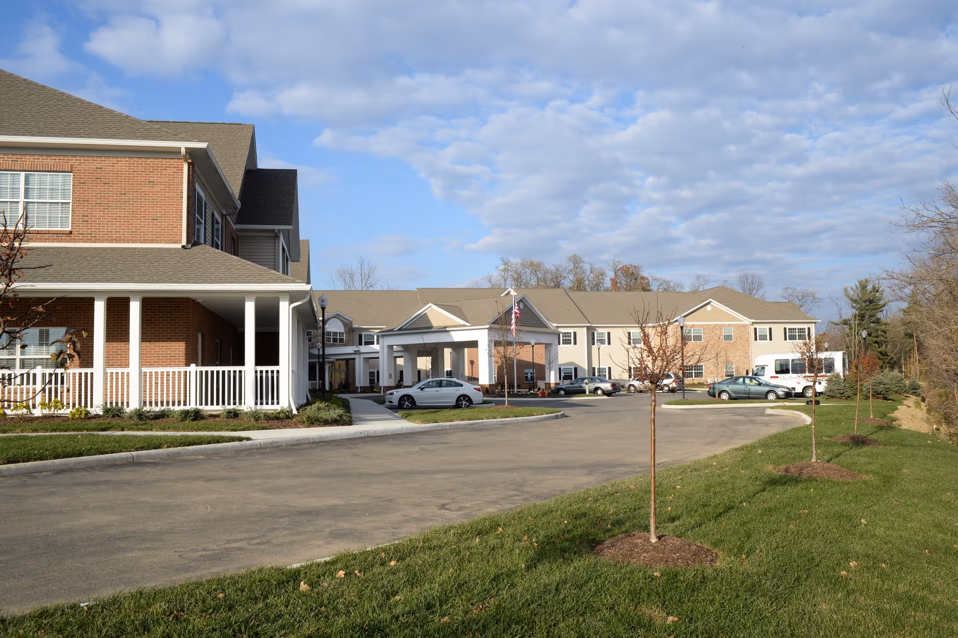 Front exterior of the Danbury Tallmadge senior living building with a covered entrance, driveway, parked cars, and landscaped grounds under a partly cloudy sky.