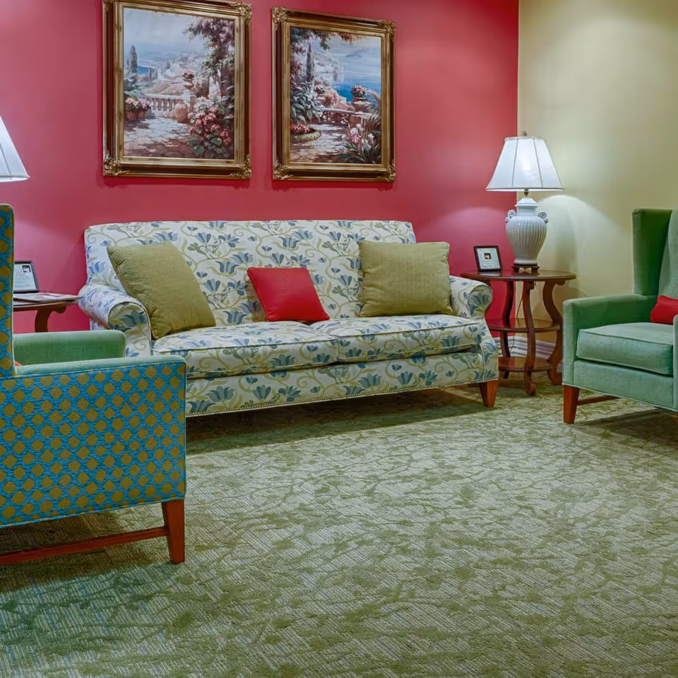 A cozy sitting area with a patterned sofa, green armchairs, side tables with lamps, and framed paintings on a red wall.