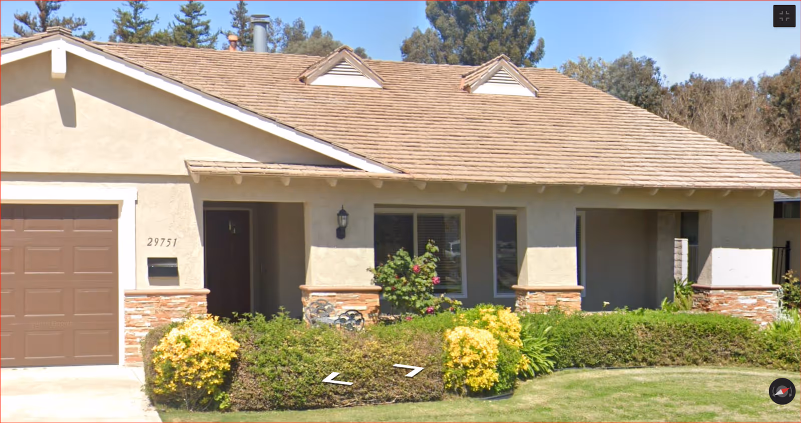 Single-story stucco house front with an attached garage, covered porch, and trimmed shrubs with yellow flowering bushes.