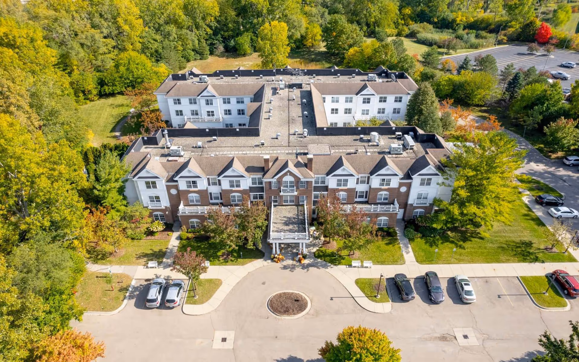 Aerial view of a three-story senior living building with a circular drive, parked cars, and surrounding trees.