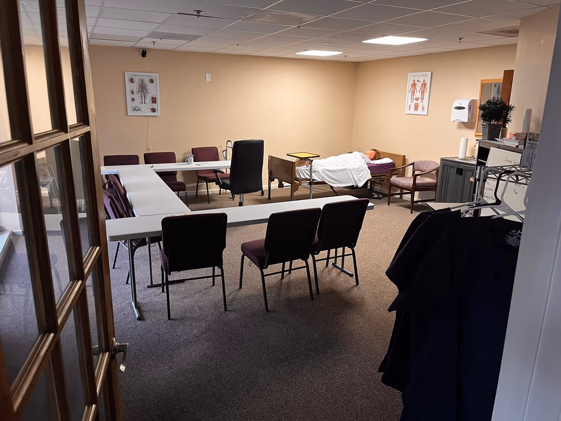 A common room with tables, chairs, and a patient lying in a bed.