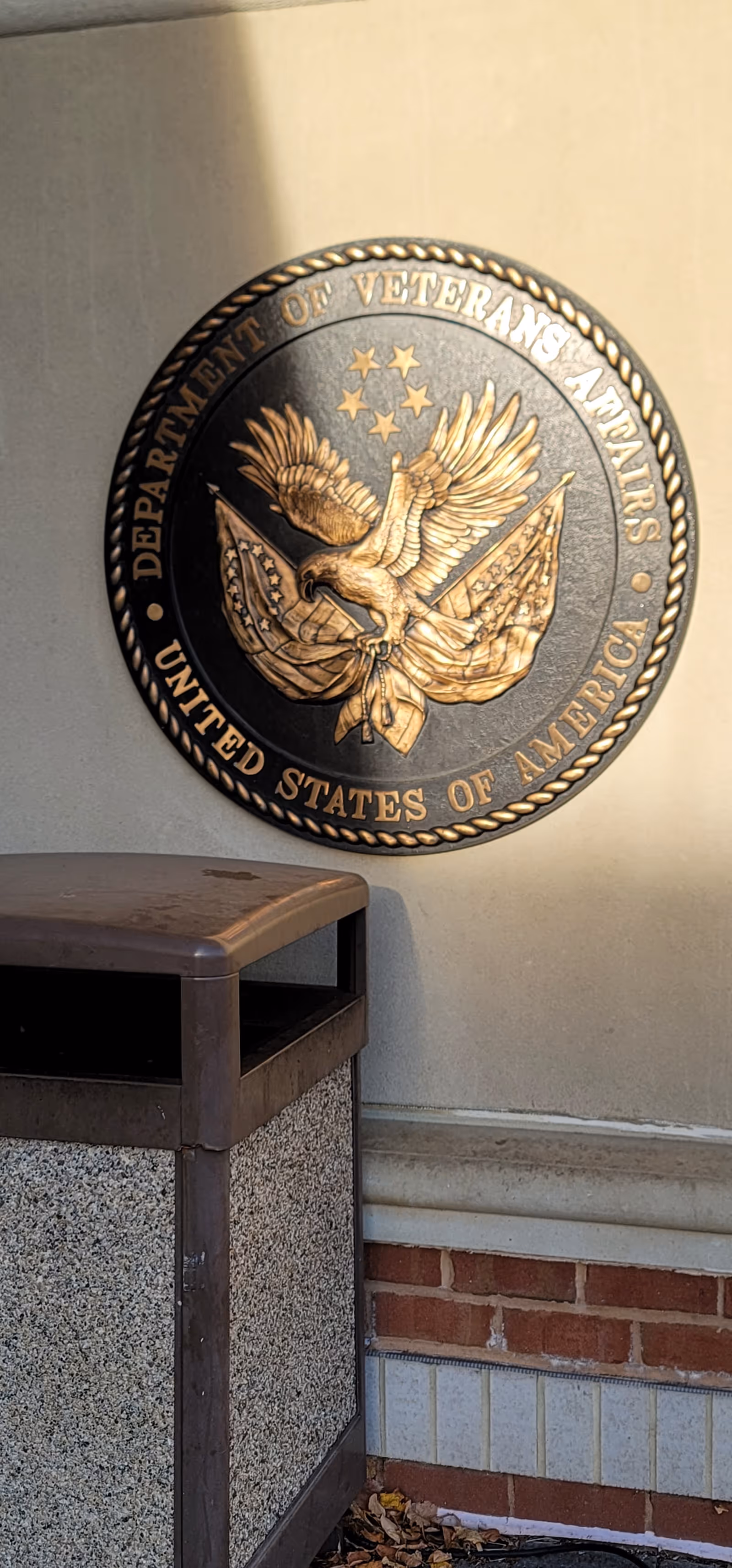 A large circular Department of Veterans Affairs plaque on a wall beside a trash receptacle.