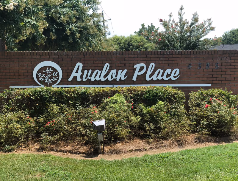 A brick wall with a white sign reading 'Avalon Place' and the number 4385, surrounded by green bushes and trees under a clear sky.