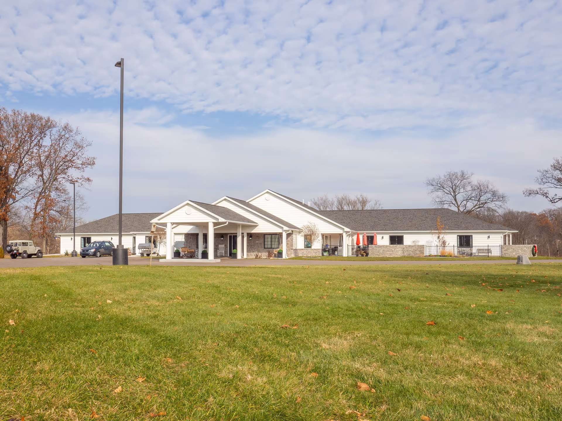 Front view of a single-story senior care building with a covered entrance, cars by the driveway and a grassy lawn in the foreground.