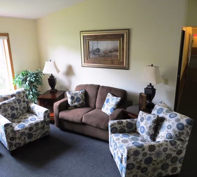 Cozy lounge area with a brown sofa, patterned chairs, lamps, and framed wall art.