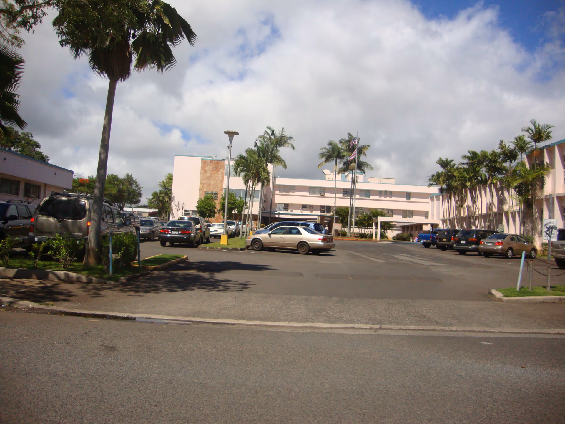 Parking lot in front of a pale pink, low-rise building with palm trees.