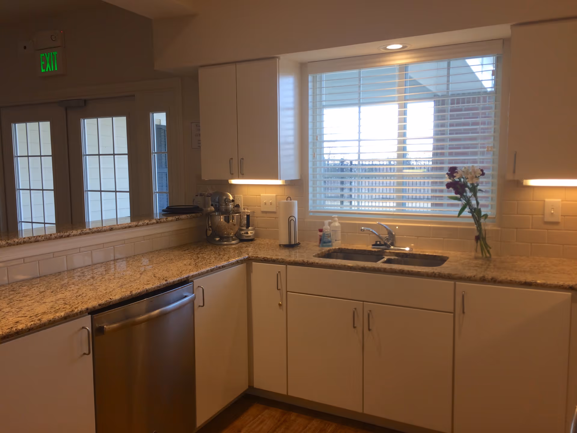 A clean kitchen area with white cabinets, granite countertops, a stainless steel dishwasher, a double sink under a window with blinds, and a vase with flowers on the counter. There is a mixer and other kitchen items on the counter, and an exit sign is visible above a set of glass doors.