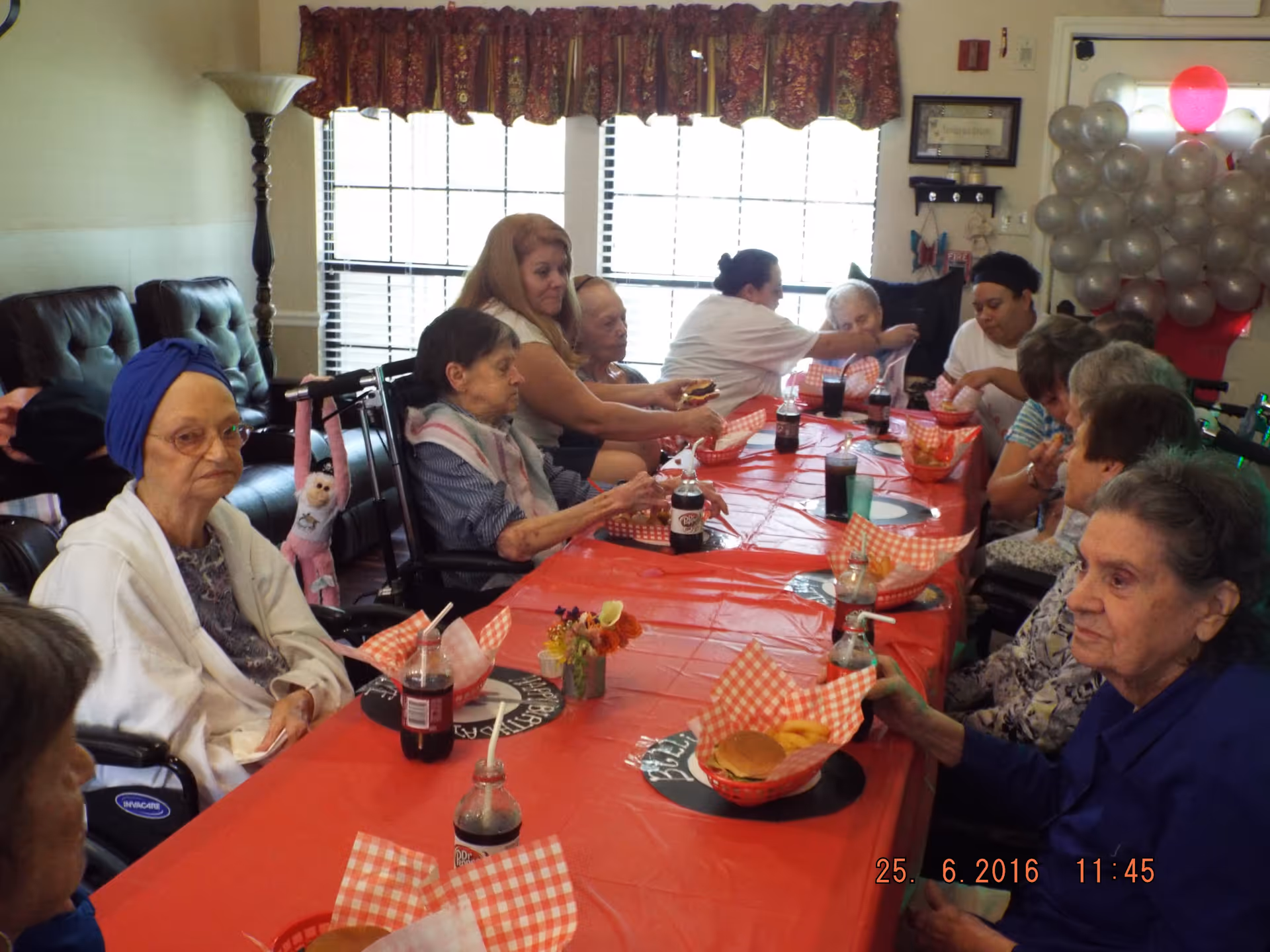 A group of elderly individuals and caregivers sitting around a long table covered with a red tablecloth, eating burgers and fries. The room has large windows with red patterned valances, a floor lamp, and a cluster of silver and red balloons near the door. Some people are in wheelchairs, and there are bottles of soda on the table.