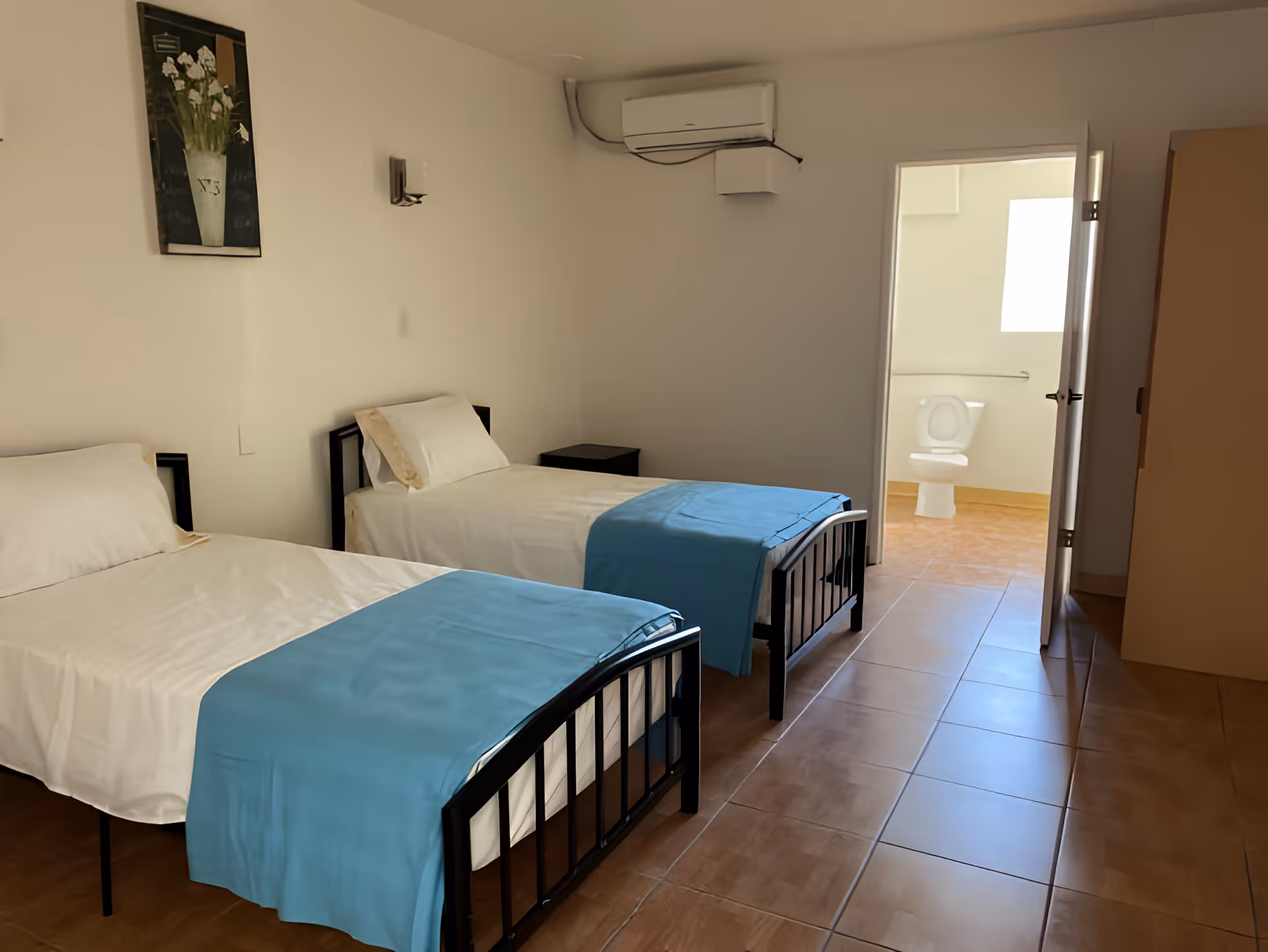 A simple bedroom with two single beds, each made with white sheets and light blue blankets at the foot. There is a small black nightstand between the beds. A painting of white flowers in a vase hangs on the wall above the beds. An air conditioning unit is mounted on the wall near the ceiling. The door to an adjoining bathroom is open, showing a toilet and a window letting in natural light. The floor is tiled in a light brown color.