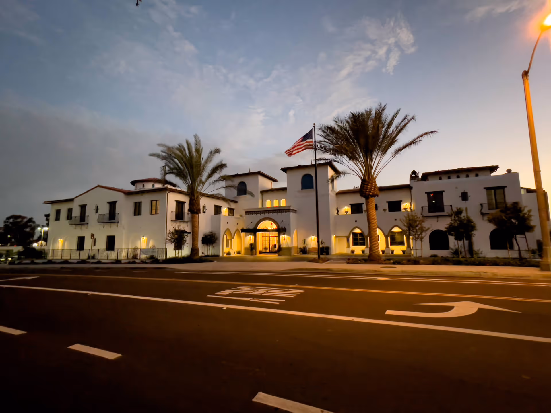 Exterior view of a large, white, two-story building with Spanish-style architecture at dusk. The building is illuminated with warm lights and features palm trees and an American flag in front. The street in front has road markings and a streetlight on the right side.