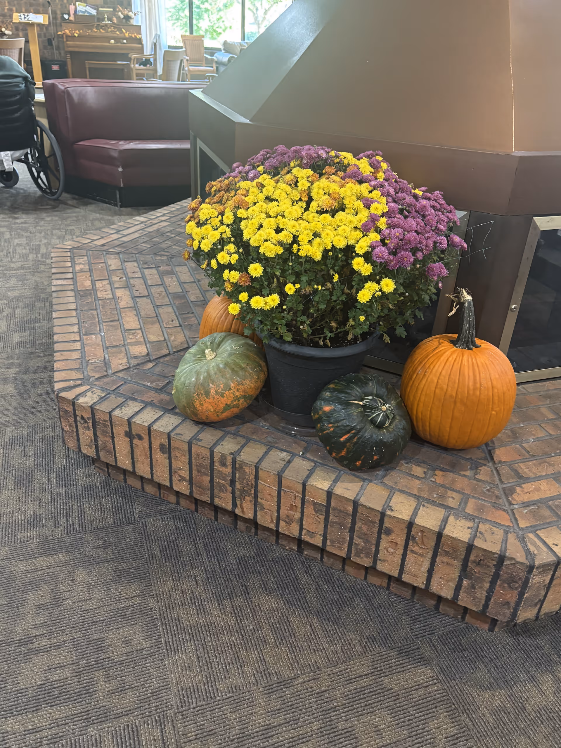 Potted yellow and purple mums and pumpkins arranged on a brick hearth in a senior living facility common room with seating and a wheelchair visible in the background.