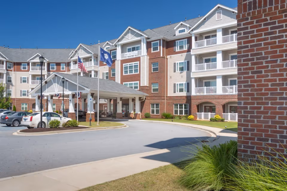Exterior view of a senior living facility named Harmony at Five Forks, showing a multi-story building with balconies, a covered entrance with flagpoles displaying the American flag and another flag, parked cars, and well-maintained landscaping under a clear blue sky.