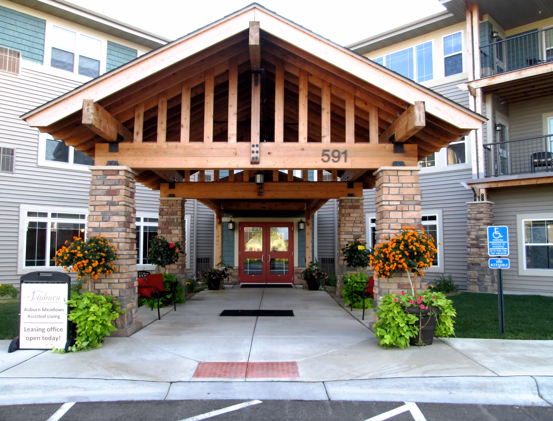 Entrance of a senior living facility with a wooden covered porch supported by brick pillars. The building has beige siding and multiple windows. There are flower pots with orange and green plants on either side of the entrance. A sign on the left reads 'Auburn Meadows Assisted Living Leasing office open today!' and a blue handicap parking sign is visible on the right.
