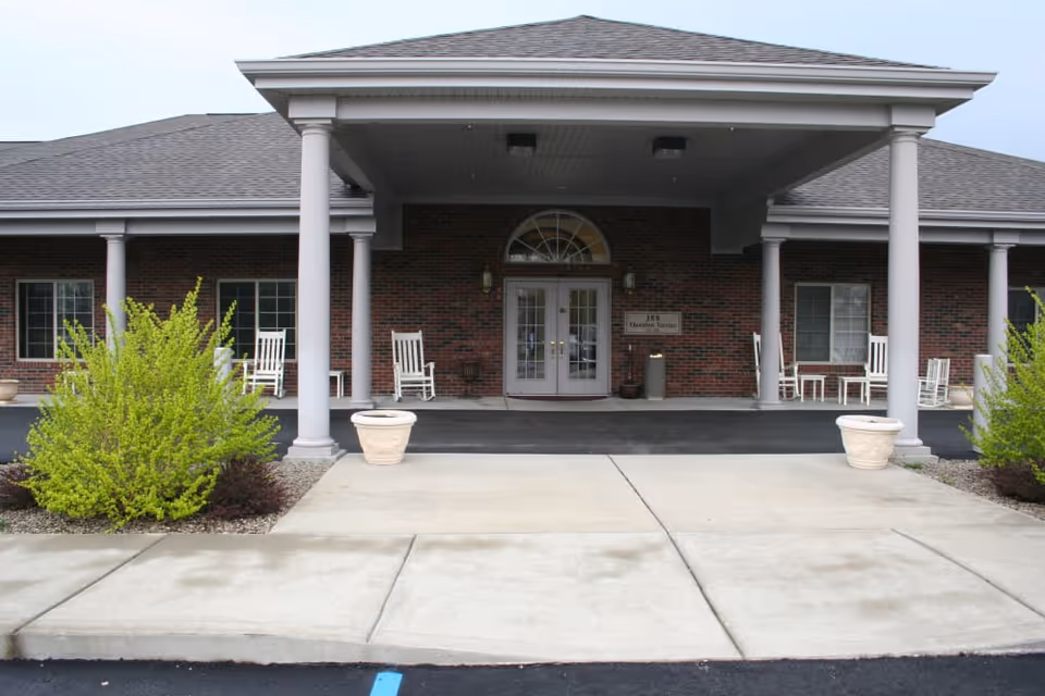Covered front entrance of a brick senior living facility with columns, rocking chairs, potted plants, and double glass doors.