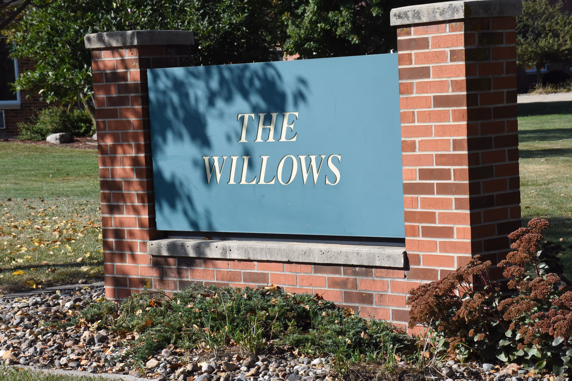 Teal sign reading The Willows between brick pillars in front of a landscaped lawn.