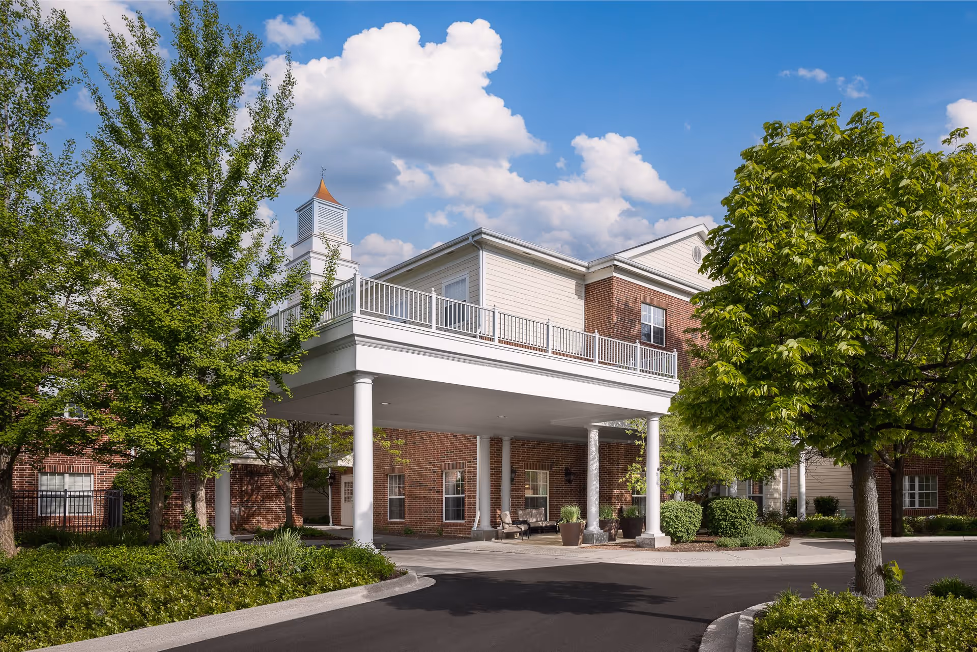 Exterior view of Belmont Village Senior Living Geneva Road showing a brick building with white trim and a covered entrance supported by white columns. The scene includes green trees and shrubs under a blue sky with scattered clouds.