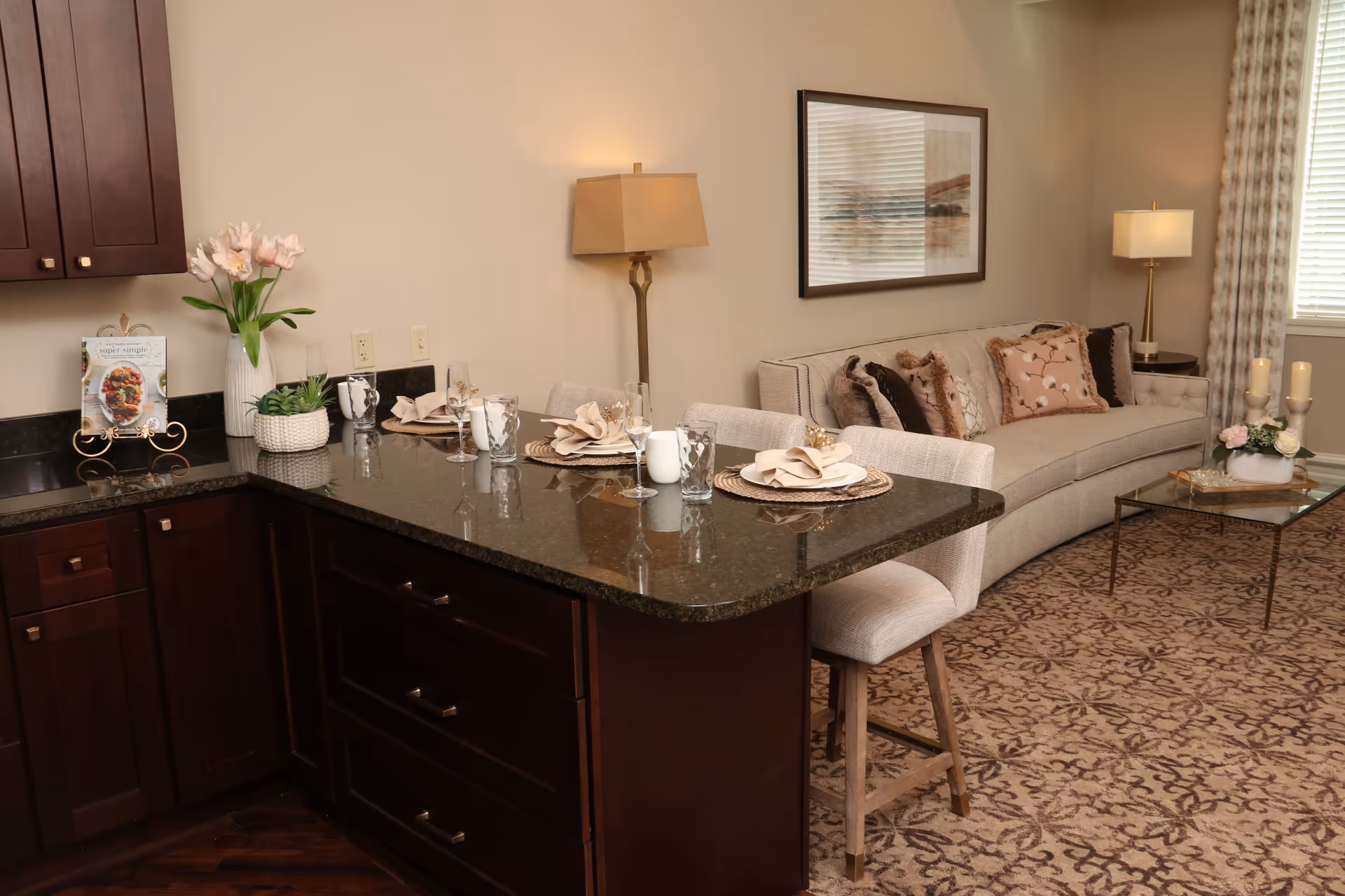 A cozy living space featuring a dark granite countertop with place settings for three, including plates, napkins, glasses, and utensils. Behind the counter is a beige sofa with multiple decorative pillows, a floor lamp, a framed picture on the wall, and a glass coffee table with candles and a floral arrangement. The room has patterned carpet and a window with blinds and curtains.