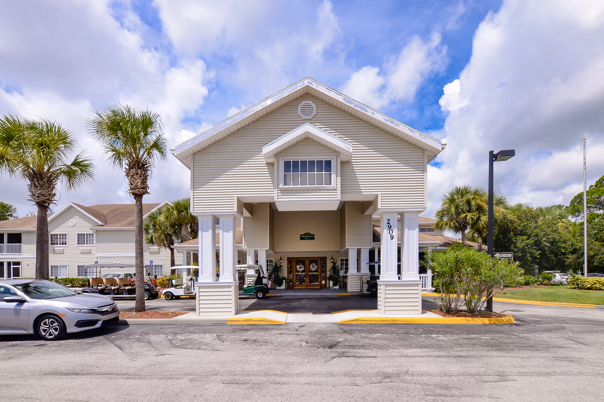 Front exterior view of a senior living facility named Lake Forest Park with a covered entrance supported by white columns, surrounded by palm trees and parked cars under a partly cloudy sky.