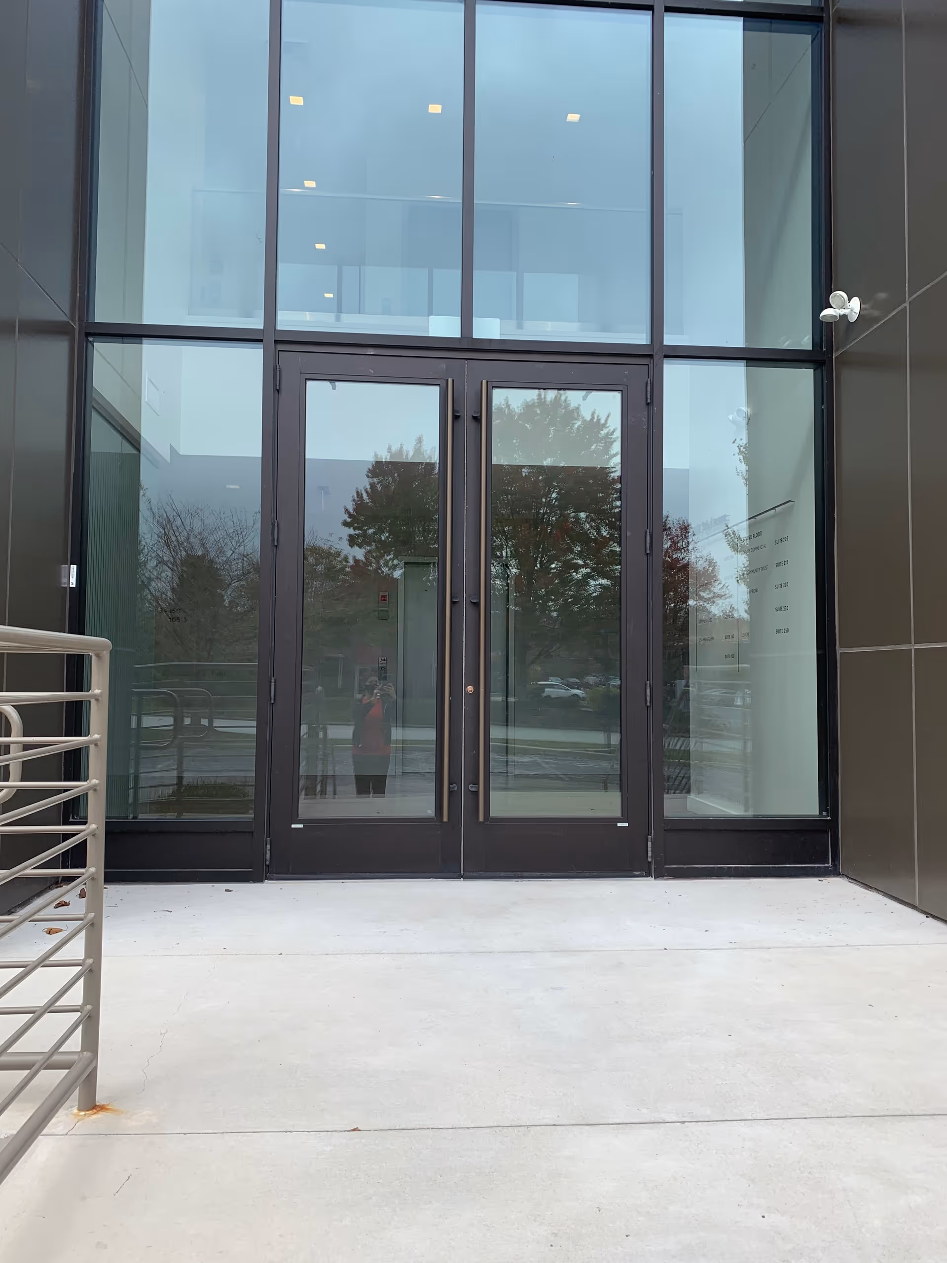 Modern glass entrance with double doors and metal frame, reflecting trees outside.