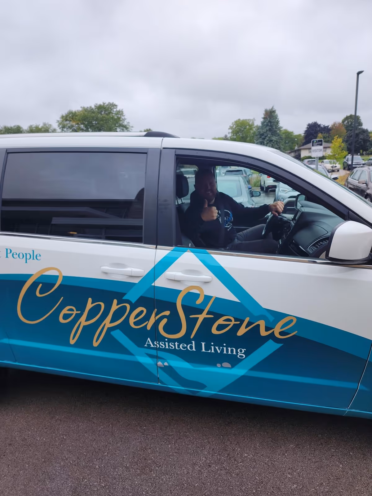 A person sitting in the driver's seat of a white and teal vehicle with the words 'CopperStone Assisted Living' on the side, giving a thumbs-up gesture. The vehicle is parked outdoors with trees and other cars in the background under a cloudy sky.