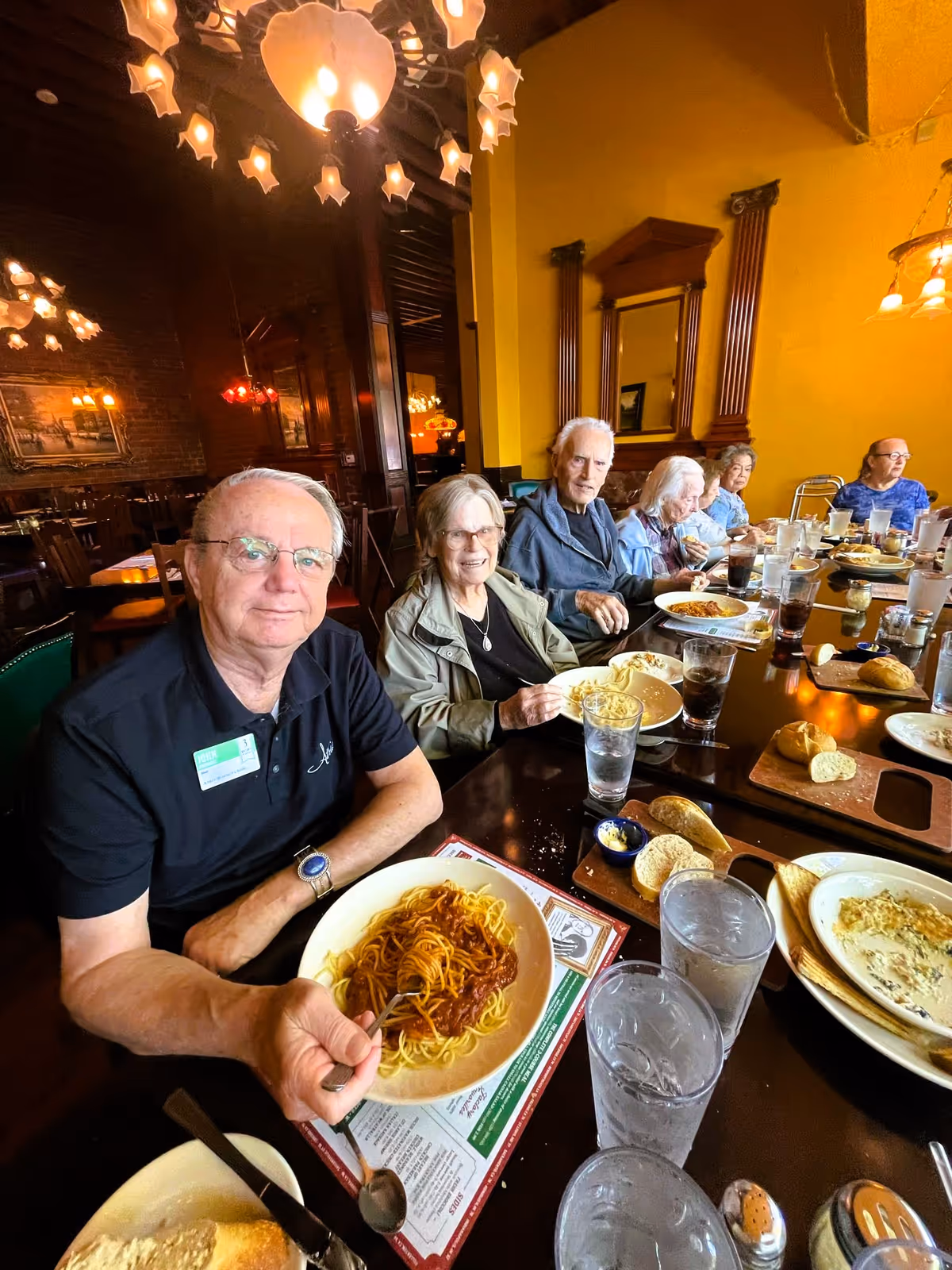 A group of elderly people sitting around a long dining table in a warmly lit restaurant, enjoying plates of food including spaghetti and bread. The setting features yellow walls, wooden accents, and decorative lighting fixtures. The people appear to be socializing and having a meal together.