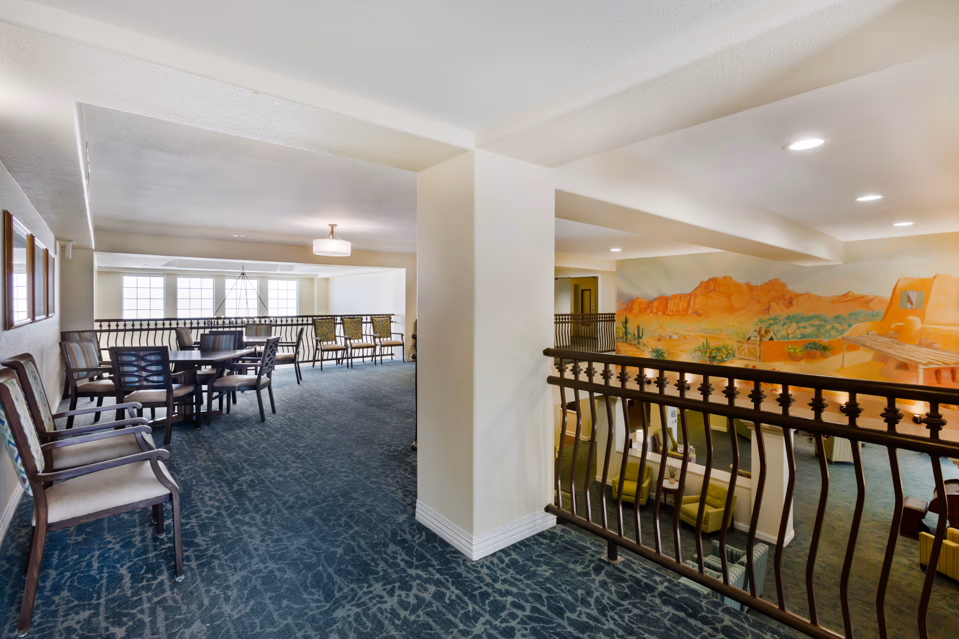 Bright interior mezzanine lounge with tables, chairs, and a desert mural overlooking the lower level.