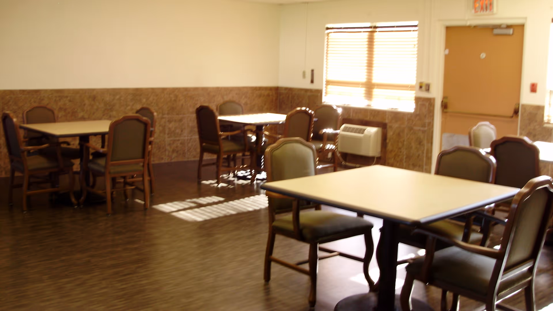 A dining area with several square tables and wooden chairs with cushioned seats and backs. The room has tiled lower walls, a window with blinds letting in sunlight, and a door with an exit sign above it.