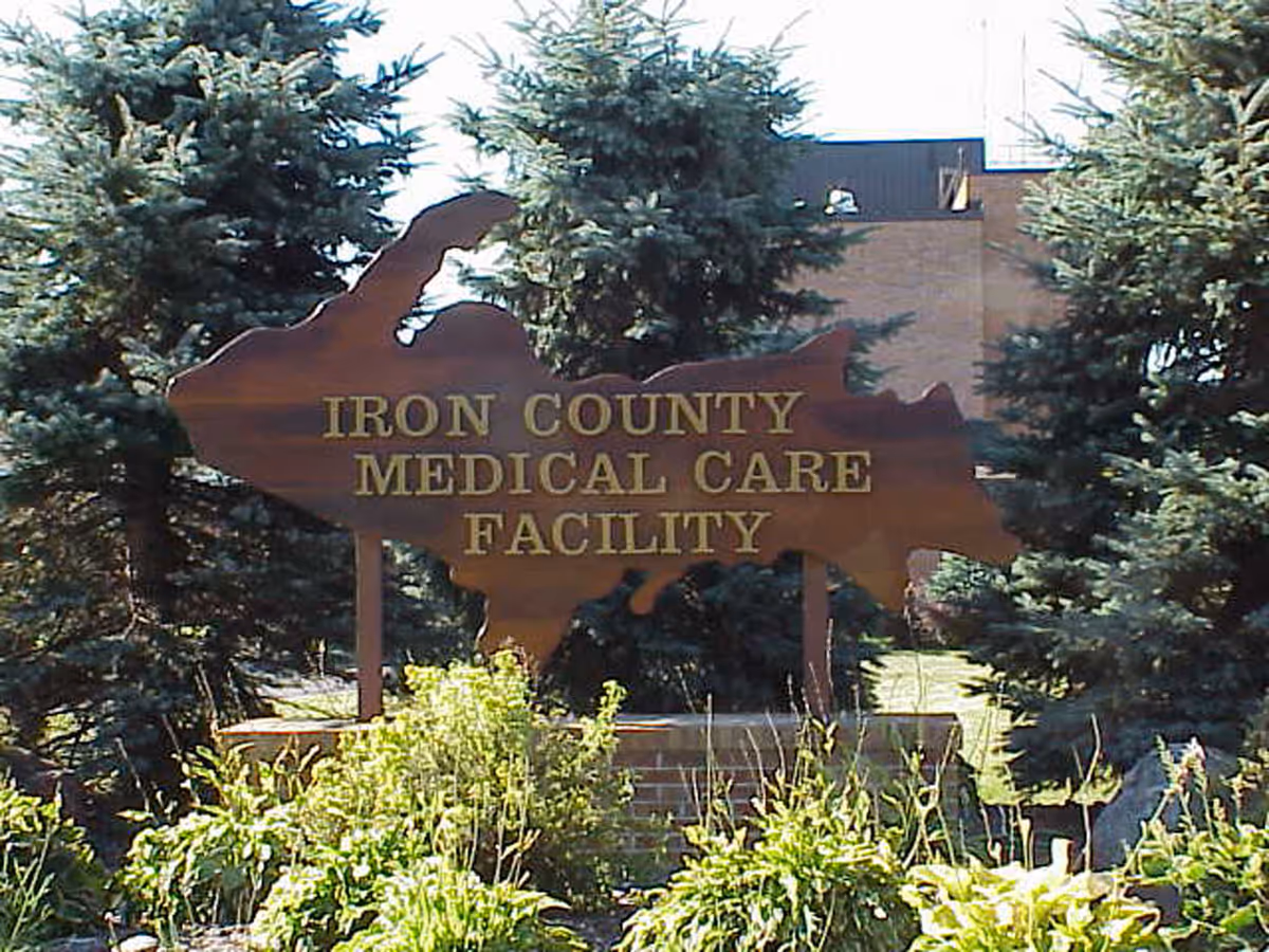 Rust-colored sign reading Iron County Medical Care Facility, framed by evergreen trees.