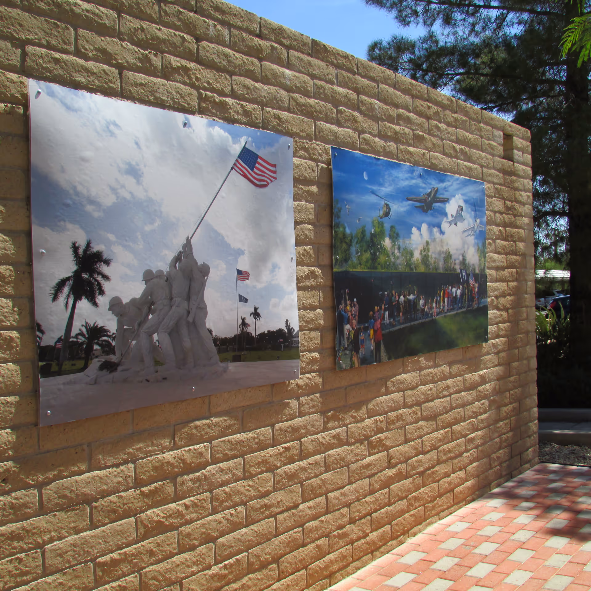 Two large photographic prints mounted on a tan brick wall outdoors. The left print shows the iconic Iwo Jima flag-raising statue with an American flag waving against a partly cloudy sky. The right print depicts a mural with military aircraft flying above a crowd of people standing in front of a black reflective wall, likely a war memorial. Trees and parked cars are visible in the background.